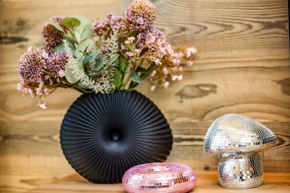 ...noch mehr Details Schwarze Vase mit bunten Trockenblumen, silberner Pilz und pinkfarbener Form auf Holzoberfläche.