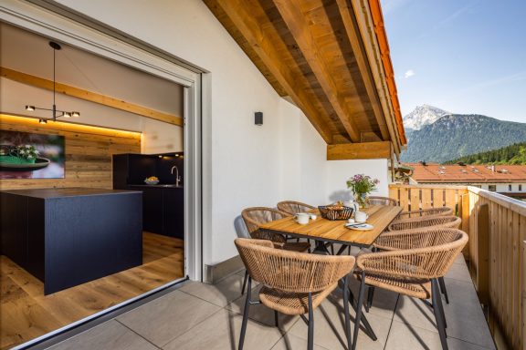 Der große Balkon lädt zum Verweilen vor der beeindruckenden Kulisse des Watzmanngipfels ein Balkon mit Holzmöbeln, Blick auf die Berge und eine offene Küche im Hintergrund.