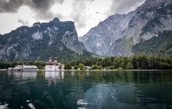 St. Bartholomä Königssee Berglandschaft mit einem See und einer kleinen, historischen Fabrik im Vordergrund.