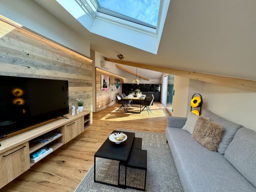 Der gemütliche Wohnbereich lädt zum Kuscheln ein Helles, modernes Wohnzimmer mit Sofa, Fernseher und Esstisch unter einem Dachfenster.